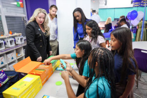 A group of students gathers around a table exploring 3D printing pens and colorful student kits, while adults observe and engage with them in a vibrant classroom equipped with 3D printers and STEM supplies.