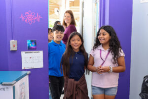A group of elementary students smiles as they walk into a brightly painted purple classroom, greeted by an adult standing at the door, with science-themed wall decals and an emergency map visible.