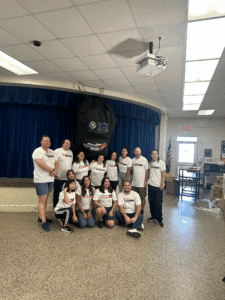 A group of Disney Cruise Line VoluntEARS from the Disney Wish poses together inside a school auditorium, standing beneath an oversized backpack labeled “Supply Zone for Teachers.” They are smiling and wearing matching white VoluntEARS shirts after helping set up for the Back-to-School Blast-Off event at Clearlake Education Center.