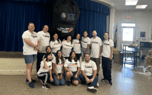 A group of Disney Cruise Line VoluntEARS from the Disney Wish poses together inside a school auditorium, standing beneath an oversized backpack labeled “Supply Zone for Teachers.” They are smiling and wearing matching white VoluntEARS shirts after helping set up for the Back-to-School Blast-Off event at Clearlake Education Center.