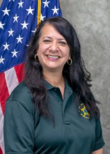 Headshot of Mindy Levy smiling in a dark green Brevard County Sheriff’s Office shirt, with an American flag and gray backdrop behind her.